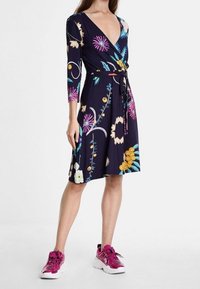 Šaty do poloviny stehen s volným střihem v námořnické modři s barevnými květinovými vzory, dlouh ými rukávy a opaskem. Kombinované s růžovými sportovními teniskami.