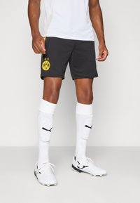 Sorte atletiske shorts med gult BVB-logo, parret med hvide sokker med sort Puma-logo og sorte og hvide fodboldstøvler.