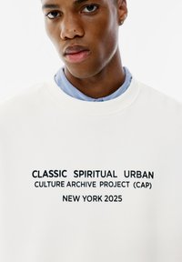 Sweatshirt bianco con testo nero: "CLASSIC SPIRITUAL URBAN CULTURE ARCHIVE PROJECT (CAP) NEW YORK 2025." Design con collo rotondo a costine.