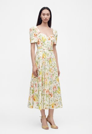 Kvinde iført en midi blomsterkjole med pufærmer, et hjerteudskæring, et tætsiddende bælte i taljen og beige højhælede sandaler.