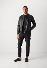 Giacca bomber nera in pelle con zip argentata, indossata sopra una maglietta nera; abbinata a pantaloni cargo scuri e zoccoli neri.