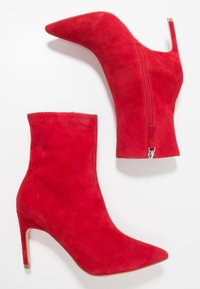 Czerwone zamszowe botki za kostkę z ostro zakończonym noskiem, wysokim obcasem typu stiletto i bocznym zamkiem. Tekstura wydaje się miękka i gładka.