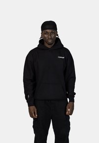 Černá mikina s kapucí a kapsou na přední straně, se symbolem "Crystal Paris" v bílé barvě. V kombinaci s černými cargo kalhotami, minimalistický design a měkká textilie.