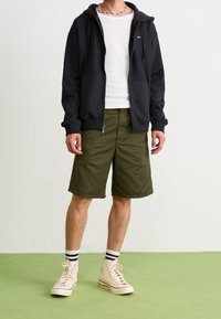 Svart zip-up hoodie, vit crew neck t-shirt, olivgröna shorts och beige sneakers med hög klack tillsammans med svarta och vita randiga strumpor.