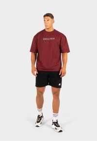 Bordeaux T-shirt met korte mouwen, voorzien van het "GORILLA WEAR" logo in grijs. Gecombineerd met zwarte shorts en witte sneakers.