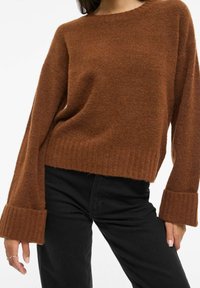 Pruun kudumiga kampsun, millel on lõdvestunud lõige, sooniline ääris ja laiad, lehvikukujulised varrukad. Sobitatud mustade kitsaste pükstega. Pehme tekstuur.