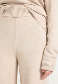 Nahaufnahme einer Person, die beiges, gestricktes Hosen und einen passenden langärmeligen Pullover mit gerippten Bündchen und Saum trägt.