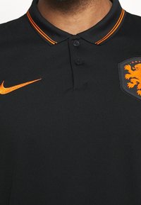 Polo nero con accenti arancioni, dotato di un'apertura con due bottoni, logo Nike ricamato e uno stemma sul lato sinistro del petto.