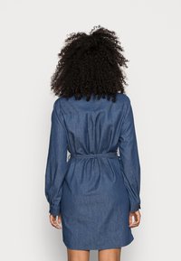 Vestido de camisa de ganga com mangas compridas, cintura justa e bainha arredondada. Apresenta uma textura suave e uma cor azul médio.