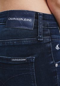 Tmavě modré džíny z denimu s viditelnou koženou náplasti s nápisem "CALVIN KLEIN JEANS" a bílými švy. Mají malé logo na zadní kapse.