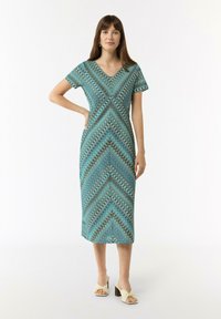 Abito a maniche corte con scollo a V in turchese, lunghezza al ginocchio, silhouette aderente, realizzato in tessuto leggero. Indossato con scarpe beige.