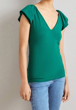 Femme portant un haut vert à col en V et manches courtes, ainsi qu'un jean bleu clair, debout devant un mur uni gris clair.