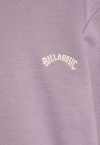 Felpa viola realizzata in un tessuto morbido, con il testo ricamato "BILLABONG" in bianco, e un design curvo nella zona del petto.
