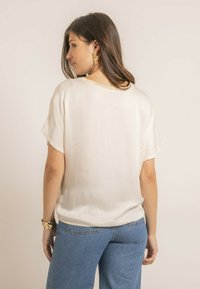 Blouse beige clair avec des manches courtes, texture lisse, col rond et un léger éclat au niveau du dos. Associée à un jean large bleu.