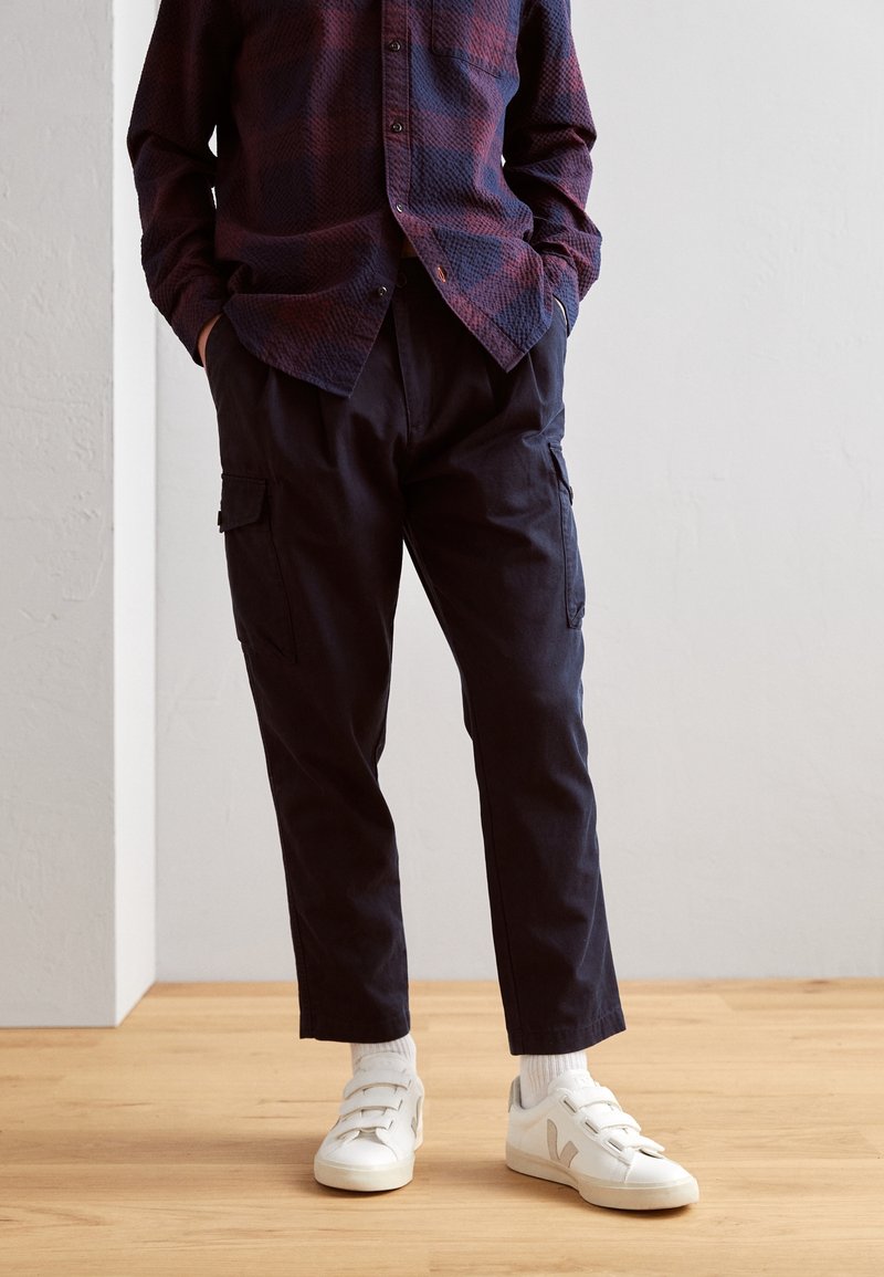 Pantaloni cargo blu navy con tasche laterali abbinati a una camicia a scacchi texturizzata blu navy e rossa. Indossati con sneaker bianche e calzini alla caviglia, su un pavimento in legno.