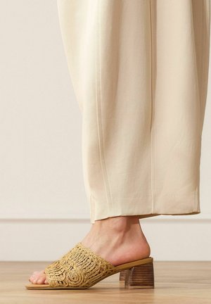 Foot wearing beige woven open-toe mule with wooden block heel, paired with loose light beige pants on a wooden floor.