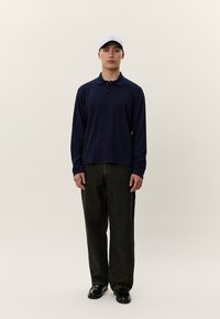 Mann som står, iført en lys grå baseballcaps, marineblå langermet polo, svarte løstsittende jeans og svarte sko mot en lys bakgrunn.