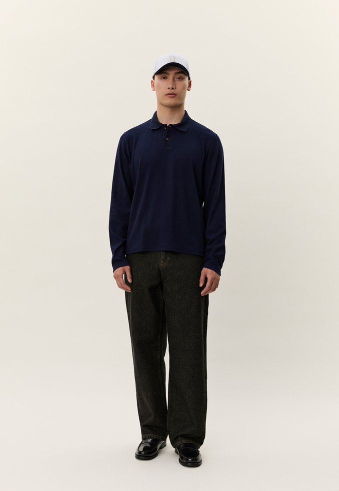 Vyras stovintis, dėvintis šviesiai pilką beisbolo kepuraitę, tamsiai mėlyną ilgomis rankovėmis polo marškinėlį, juodas laisvas džinsas ir juodus batus prieš šviesų foną.