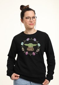 Sudadera negra con un gráfico de un personaje ilustrado rodeado de huevos de Pascua de colores. El diseño incluye tonos pastel y patrones.