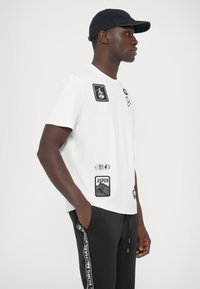Balta īsām piedurknēm T-krekls ar melnām grafiskām uzlīmēm un izšūtiem dizainparaugiem. Valkāts kopā ar melnām biksēm ar logotipa svītru sānos.