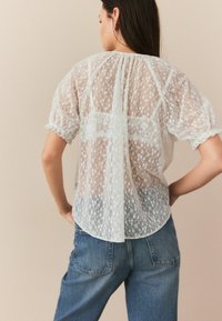 Blouse en dentelle blanche avec de courtes manches bouffantes, un col froncé et un motif floral transparent. Associée à un jean en denim bleu décontracté.