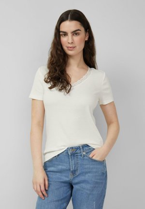 Młoda kobieta z długimi kręconymi włosami, ubrana w biały krótki top z koronkowym wykończeniem i niebieskie dżinsy, stojąca na tle szarego tła.