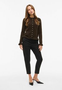 Tumši brūns kardigāns ar viļņotu apakšmalu un piedurknēm, uz kura ir dekoratīvi pogas priekšpusē. Saskaņots ar šaurām melnām biksēm un smailiem papēžiem.