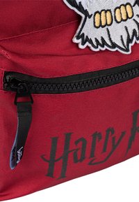 Zaino in tessuto rosso con tasca con zip, cerniera nera e toppa decorativa ricamata bianca e gialla con design grafico.