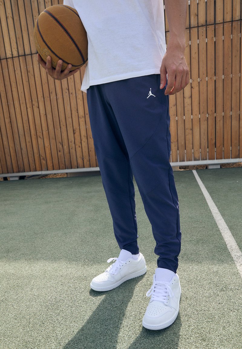 Námořnické sportovní tepláky s bílým logem Jumpman, v kombinaci s bílými nízkými teniskami. Pozadí tvoří dřevěný plot a basketbalové hřiště.