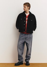 Mladi moški, oblečen v črno jakno s šerpasto ovratnico, rdečo jopa z belim potiskom, ohlapne izprane kavbojke in črne škornje, stoji ob enostavni steni.