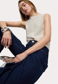 Top senza maniche beige testurizzato abbinato a jeans blu scuro. Modella con sandali e bracciale argentati, su uno sfondo semplice.