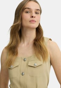 Top senza maniche beige con scollatura rotonda, dettagli con bottoni argento e due tasche frontali. Tessuto liscio con una leggera lucentezza.