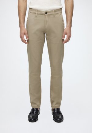 Homme portant un pantalon beige slim et des mocassins en cuir noir, debout sur un fond blanc uni.