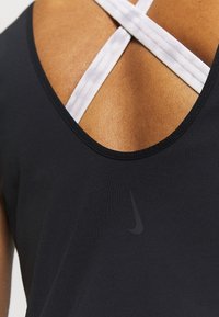 Černý sportovní top na ramínka s kulatým výstřihem a křížovými bílými pásky na zadní straně. Obsahuje jemné černé logo Nike.