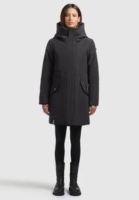 Mustast pehmendatud parka kõrge kraega, ees oleva nööpidega sulgemise ja kahe suure küljetaskuga. Materjal tundub sile ja veekindel.