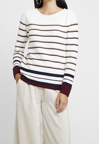 Sweter w paski w białym kolorze z pasami w granatowym, taupe i burgundowym. Z okrągłym dekoltem i prążkowanymi mankietami, zestawiony z kremowymi spodniami o szerokich nogawkach.