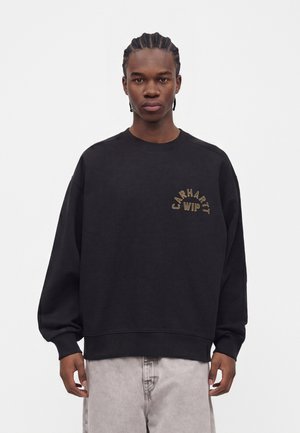 Joven con el pelo trenzado, vestido con una sudadera negra Carhartt WIP y pantalones gris claro, de pie frente a un fondo simple.