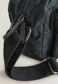Sort stof tas med lynlås, der har en tekstureret rem og metalhardware detaljer. Kompakt design med et robust udseende.
