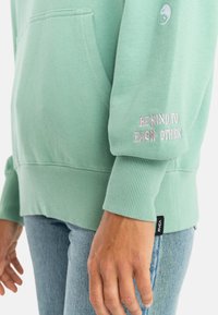 Mint green hoodie with ribbed cuffs, large pocket, and embroidered "BE KIND TO EACH OTHER" on the sleeve. Cotton material, relaxed fit.