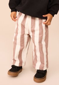 Bambino che indossa pantaloni beige e bianchi a righe verticali, felpa nera e scarpe nere con suole marroni, in piedi su un pavimento beige.