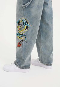 Ljust blå denimbyxor har en detaljerad broderad drakdesign i livliga färger på vänster ben, tillsammans med vita sneakers.