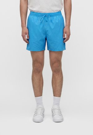 Mann trägt hellblaue Shorts mit Kordelzug, weiße Kniesocken, weiße Turnschuhe und ein weißes T-Shirt und steht vor einem einfarbigen Hintergrund.