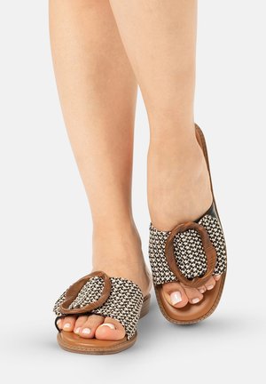 Pies con sandalias sin cierre con tiras tejidas en blanco y negro y grandes hebillas ovaladas marrones sobre suelas color tostado sobre fondo blanco.