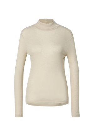 Femme debout portant un pull à col roulé beige, un jean large bleu clair et des chaussures noires, sur un fond gris uni.