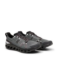 Chaussures de sport grises avec une tige en mesh, des zones renforcées et une semelle noire texturée comportant des découpes circulaires. Accents réfléchissants sur les côtés.