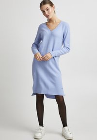 Abito in maglia azzurro chiaro con scollo a V e maniche lunghe. Presenta una superficie texturizzata, un orlo posteriore leggermente più lungo e un dettaglio con logo sul lato inferiore.