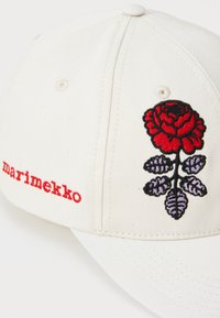 Biela bavlnená baseballová čiapka s červeným vyšitým dizajnom ruže a fialovými listami. Na prednej strane je logo "marimekko" v červenom písmene.
