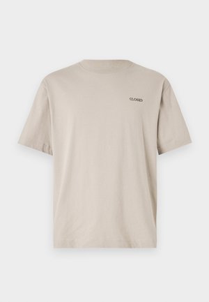 Beige miesten puuvillainen t-paita lyhyillä hihoilla, jossa on "CLOSED" logo vasemmassa rinnassa. yhdistettynä tummansinisiin housuihin ja valkoisiin lenkkareihin.