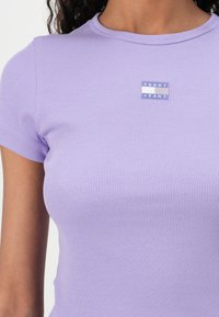 T-shirt lavande à côtes avec des manches courtes et un col rond. Présente une petite étiquette en tissu bleue avec "TOMMY JEANS" brodée à l'avant.