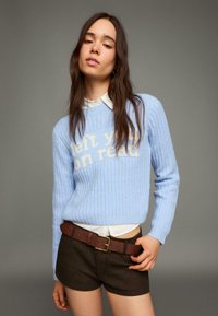 Svetlomodrý rebrovaný sveter s textom "left you on read" v krémovej farbe, nosený cez bielu košeľu s golierom, skombinovaný s hnedými šortkami a opaskom.